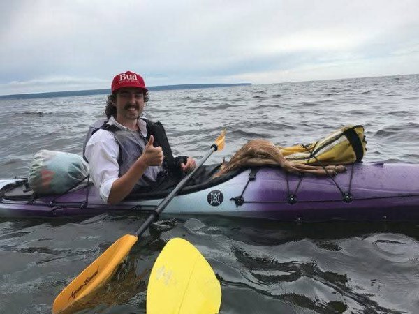 Person in a red hat kayaking, gives thumbs up on a calm body of water.